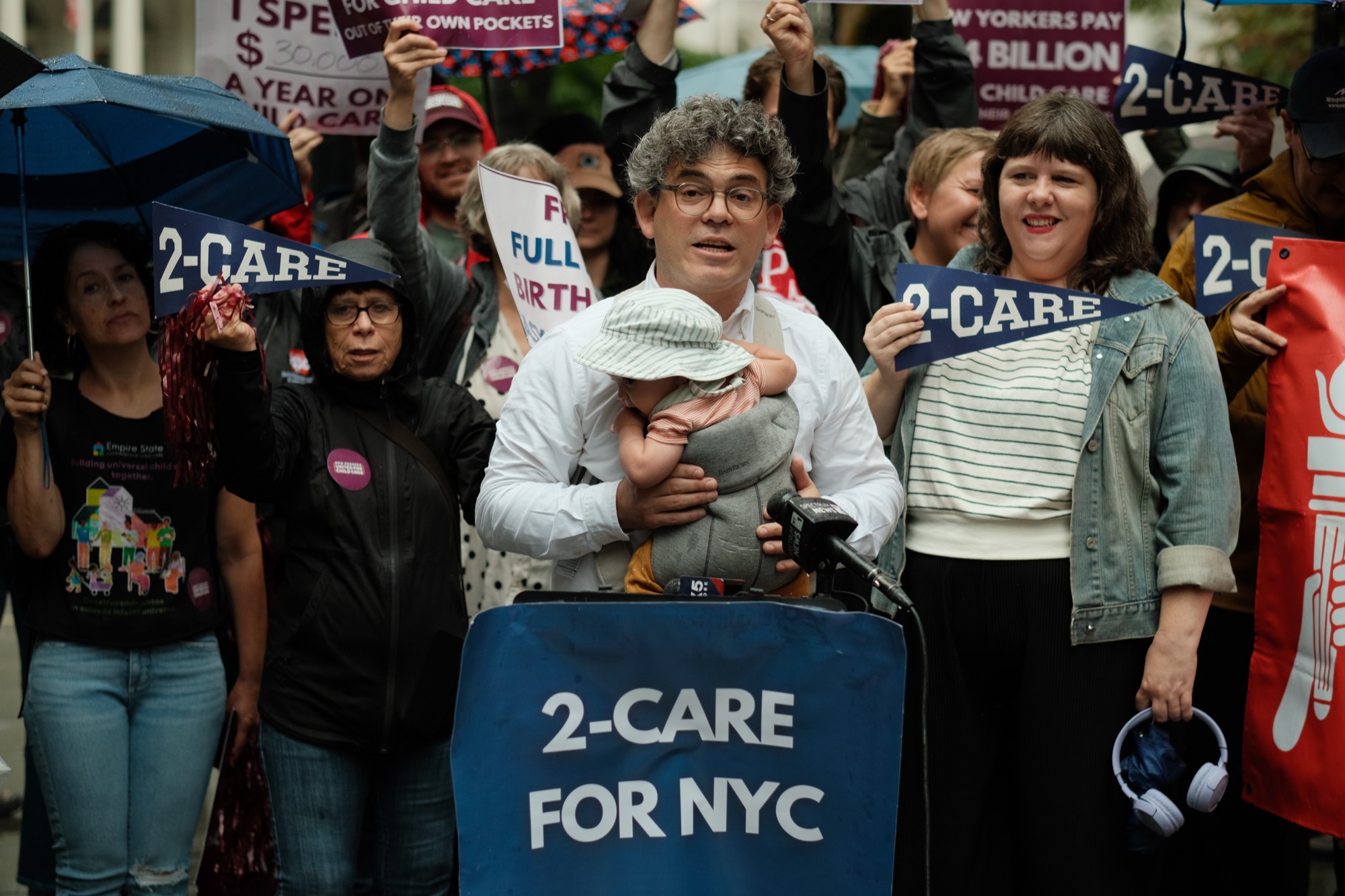 A parent holds his baby while speaking at a New Yorkers United for Child Care rally behind a “2-Care for NYC” podium, supporters and press behind him.
