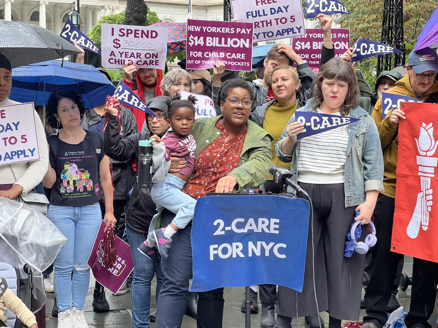 A parent holds a child at a New Yorkers United for Child Care rally behind a “2-Care for NYC” banner.