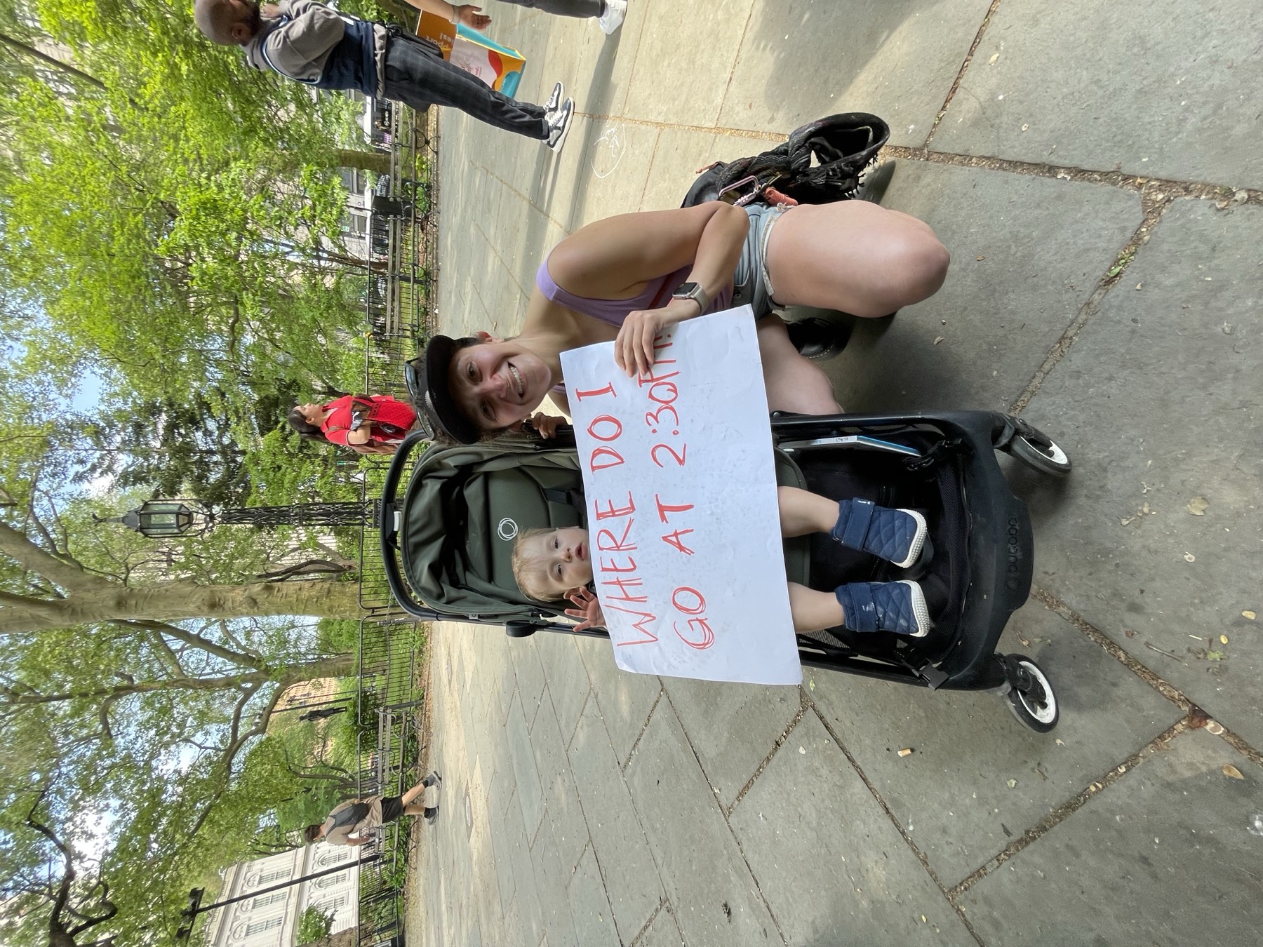 A parent kneels beside a stroller holding a hand-lettered “Where do I go at 2:30?” sign at a New Yorkers United for Child Care rally.
