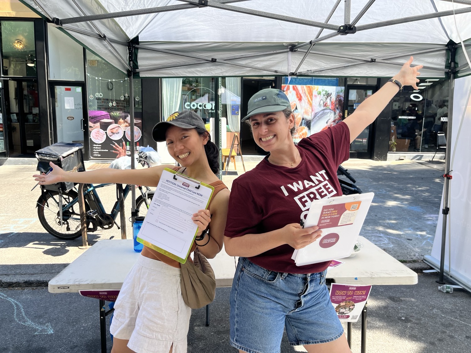 Two volunteers cheer behind a sidewalk petition table stacked with New Yorkers United for Child Care clipboards and flyers.