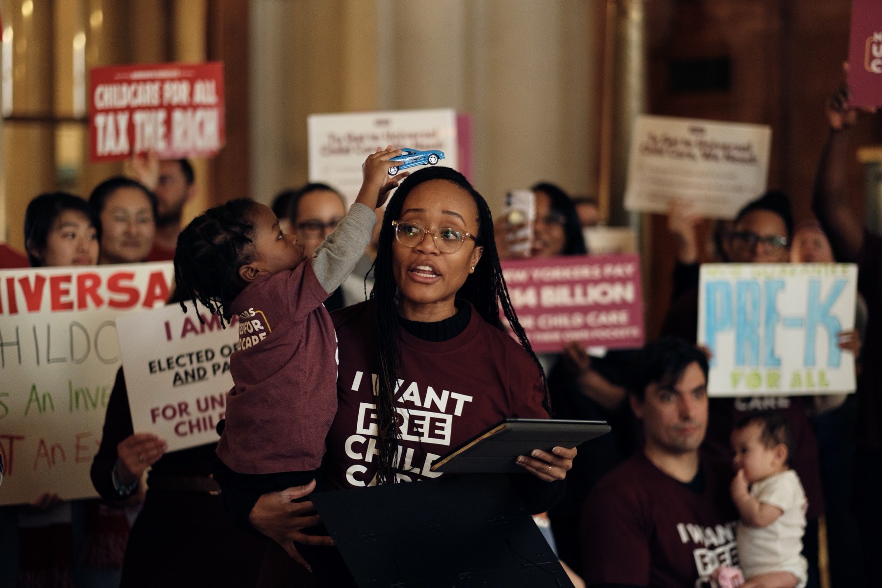 A parent in a maroon “I want free child care” shirt holds her toddler at a New Yorkers United for Child Care rally, surrounded by members and signs.