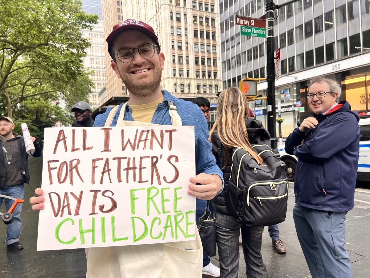 A smiling dad in a New Yorkers United for Child Care cap holds a hand-painted “All I want for Father’s Day is free child care” sign on a downtown street.